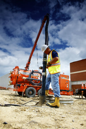 Un hombre en botas de trabajo y chaleco de seguridad de pie en un sitio en construcción con un camión con aspiradora ditch witch anaranjado